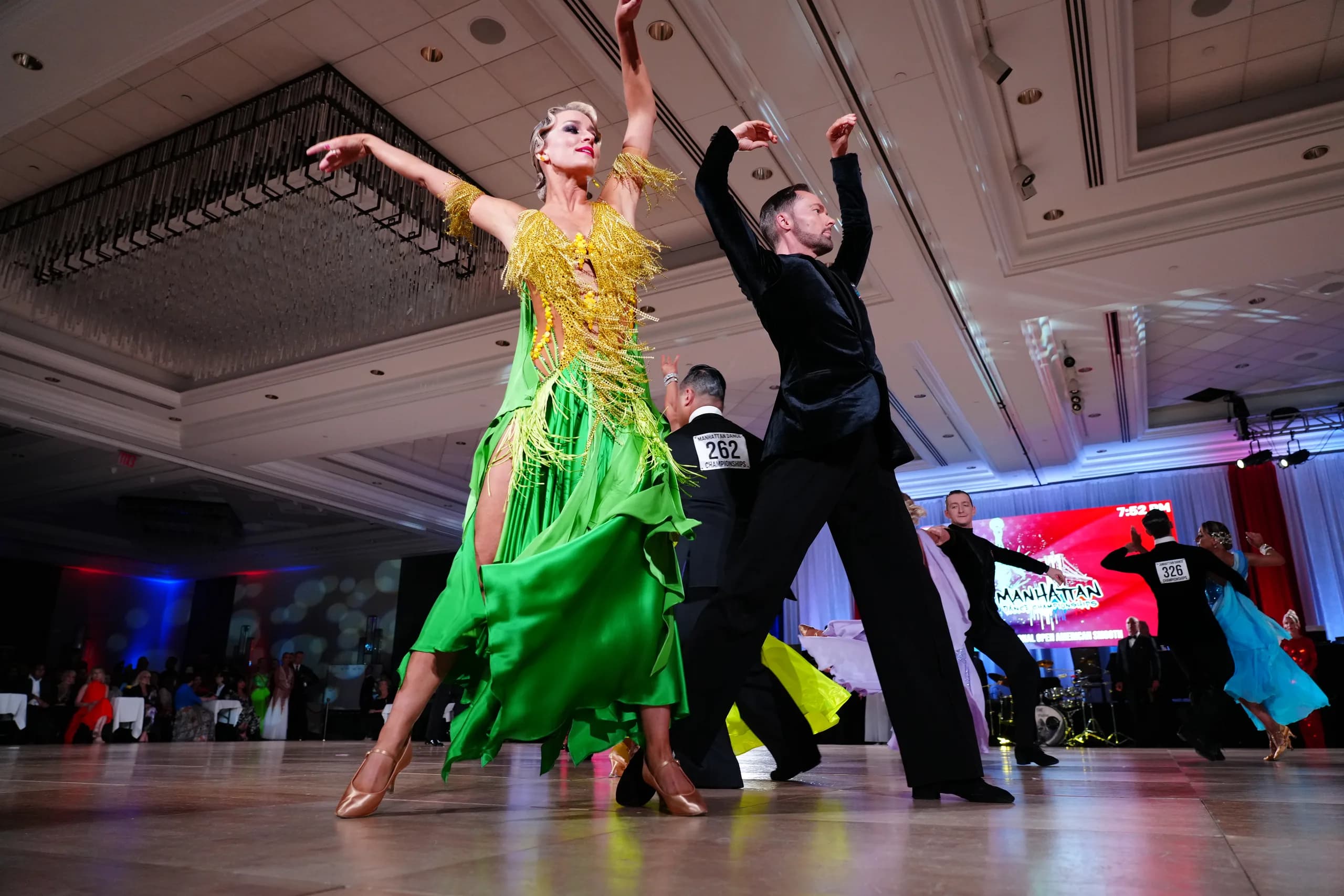 Jaimee arches in a fringed gown beside Travis under the chandeliers of a grand ballroom